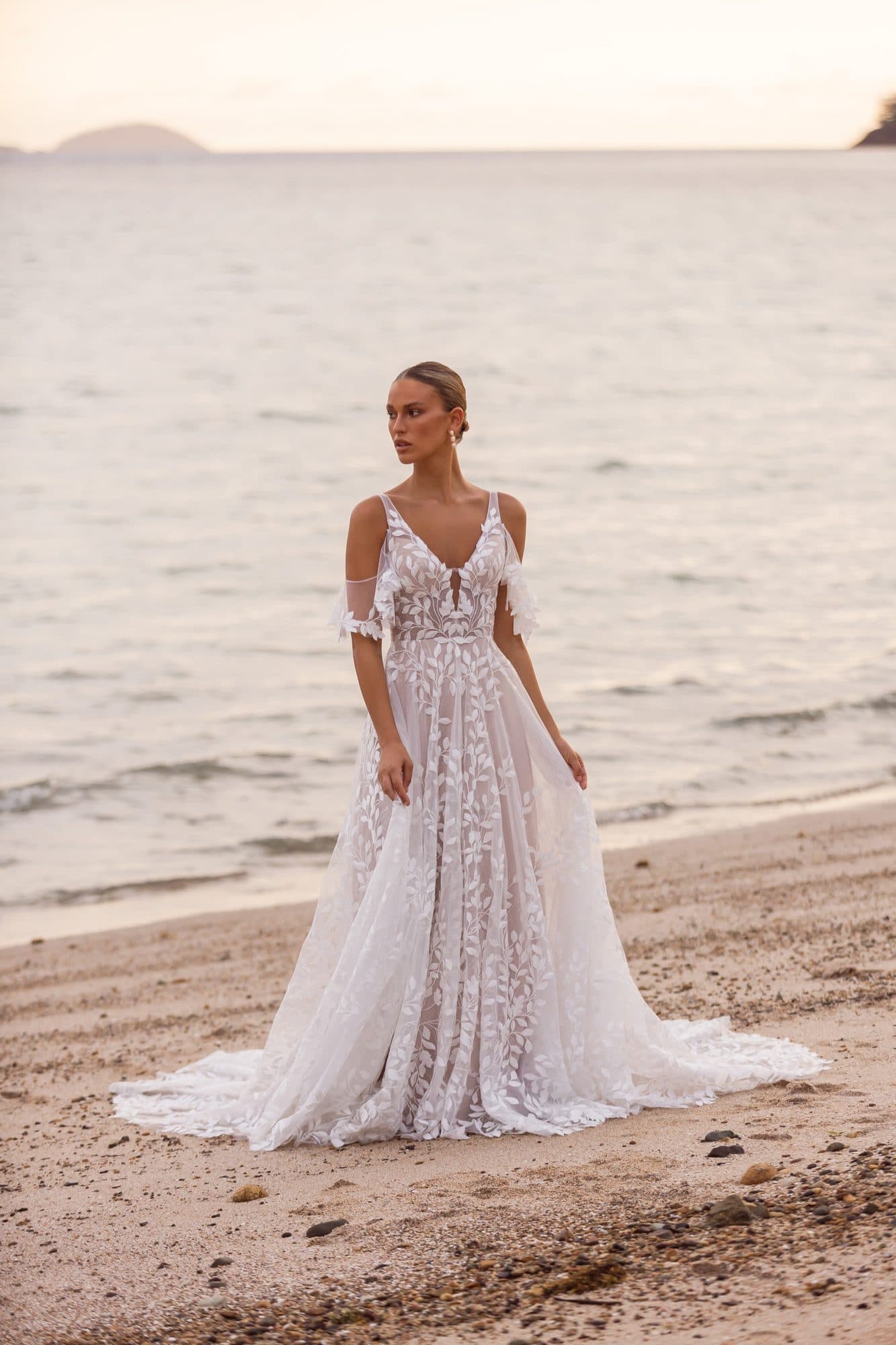Front of the Madi Lane Jaya gown, a floral lace A-line with detached flutter sleeves, worn on a beach.