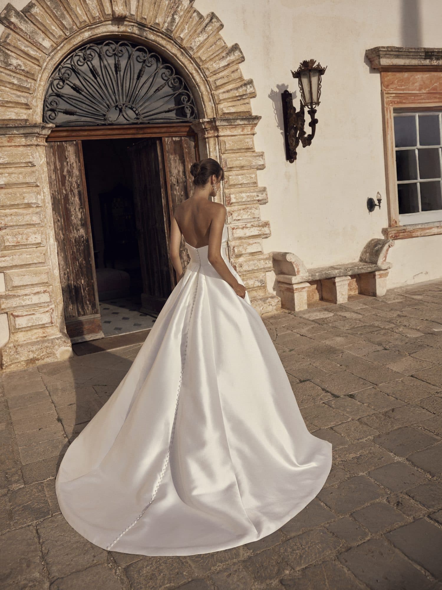 Back of the Modeca Marcella gown showing its low back and dramatic satin train at an arched doorway.