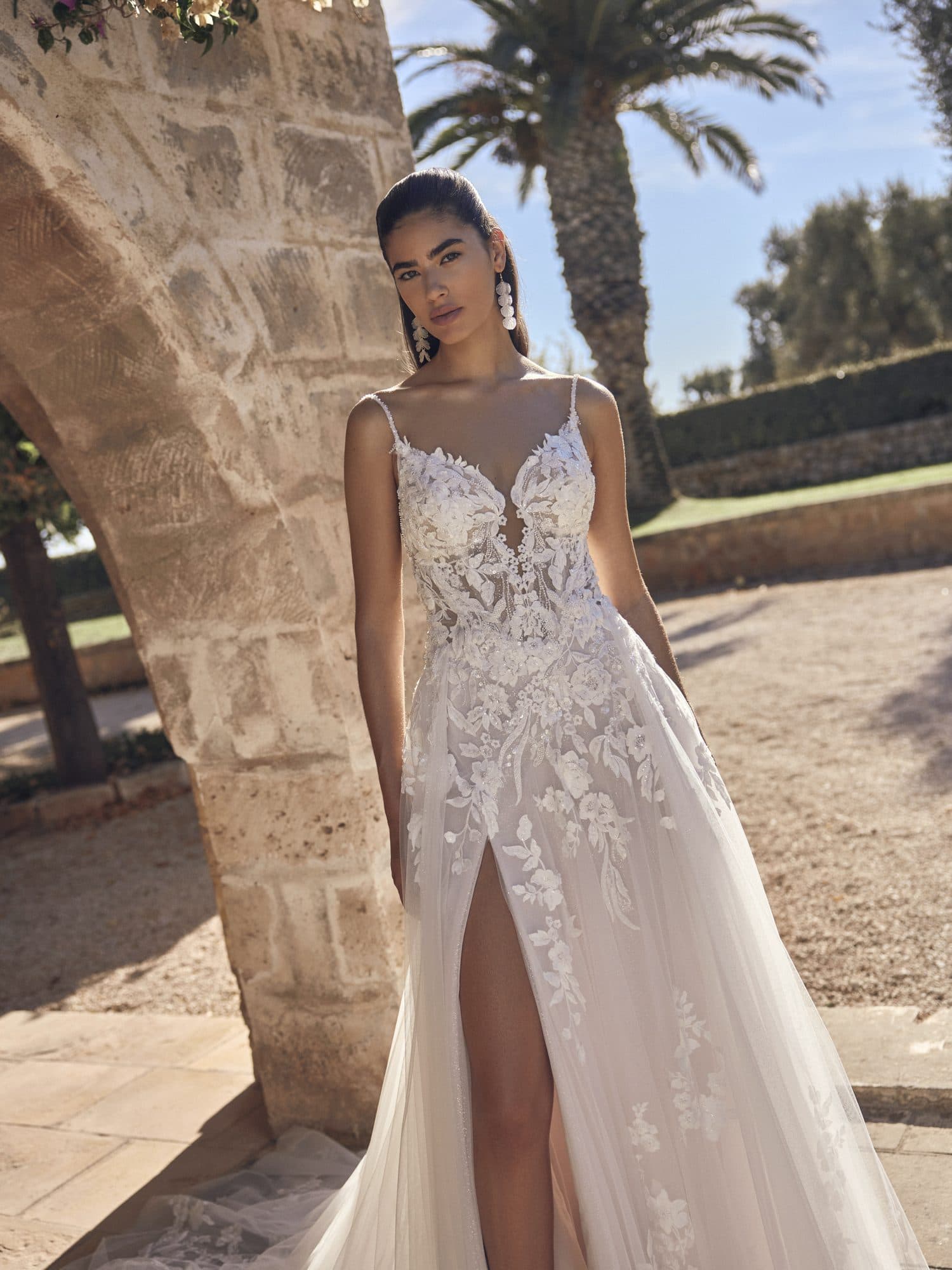 Front of the Modeca Mivonne gown, a floral lace A-line with fine straps and a leg slit, under a stone arch.