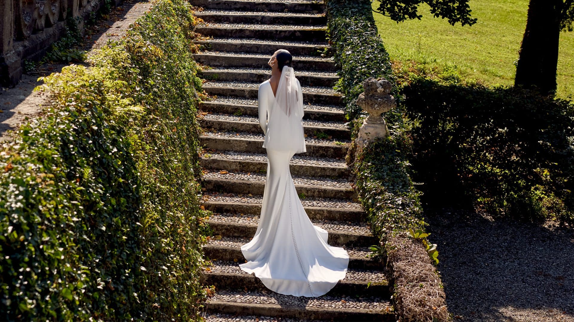 Bride seen from behind on ivy-lined stone garden steps, the train of her crepe gown flowing down the steps behind her.