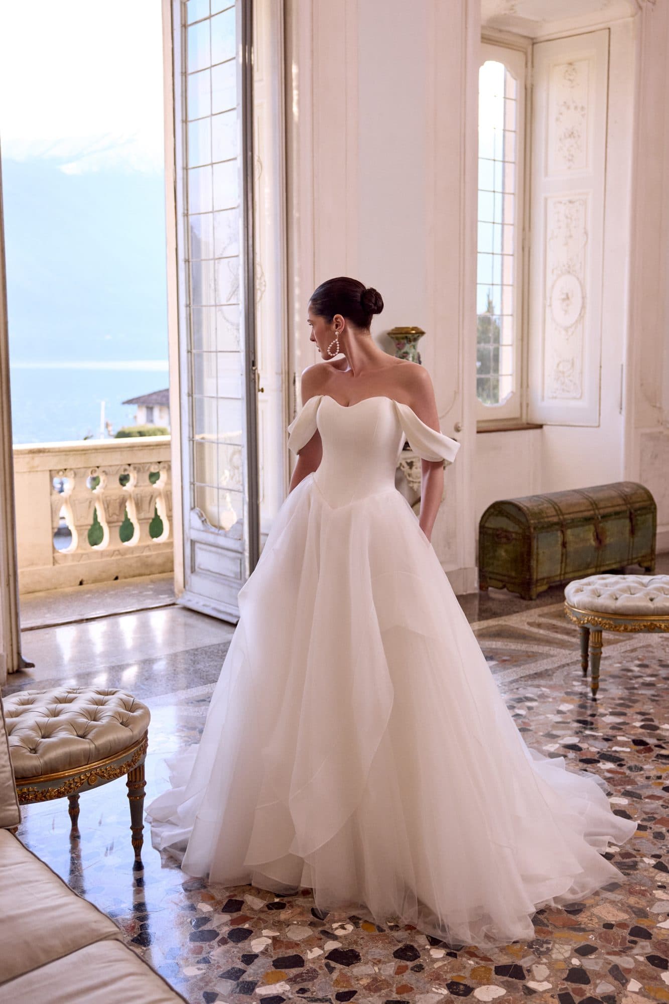 Bride in an off-the-shoulder Justin Alexander ballgown with a layered tulle skirt, standing in a lakeside villa doorway.