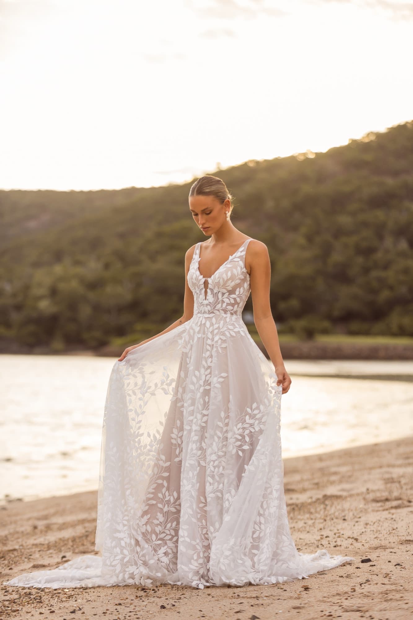 Bride in a botanical lace gown lifting her skirt as she walks along a shoreline at golden hour.