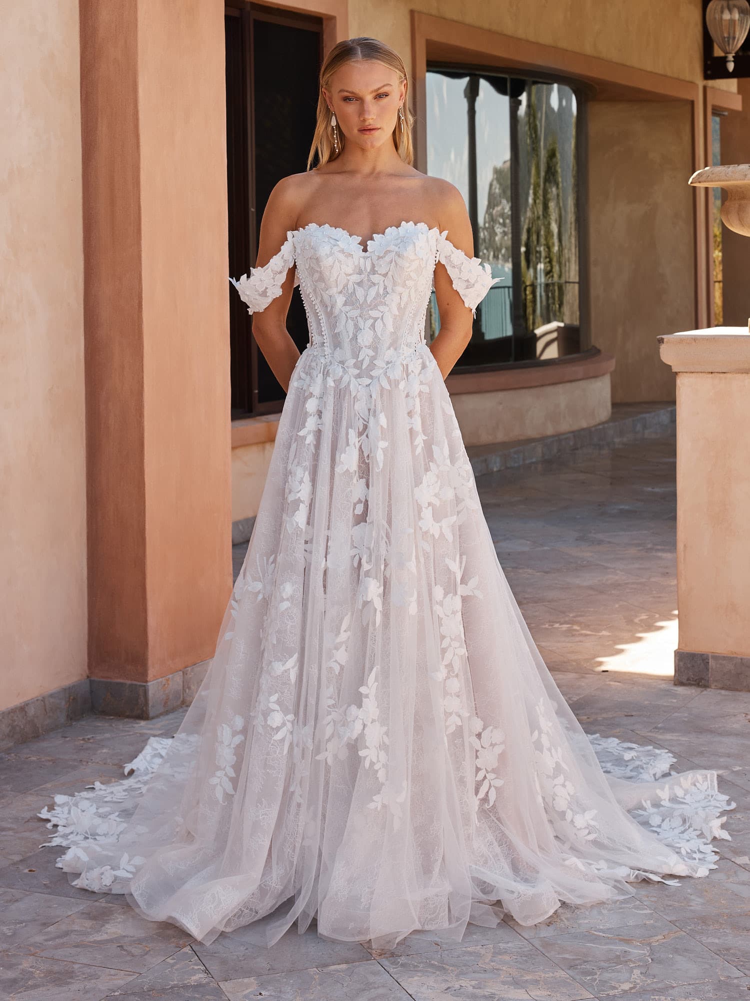 Bride in an off-the-shoulder Madi Lane gown covered in three-dimensional floral lace, standing in a sunlit courtyard.